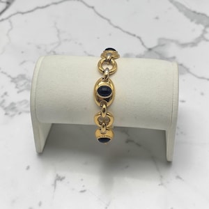 May include: Gold-toned bracelet featuring oval and circular links, each adorned with a dark blue stone. The bracelet is displayed on a white jewelry stand, showcasing its design and craftsmanship. The bracelet's links are connected by smaller, circular links.