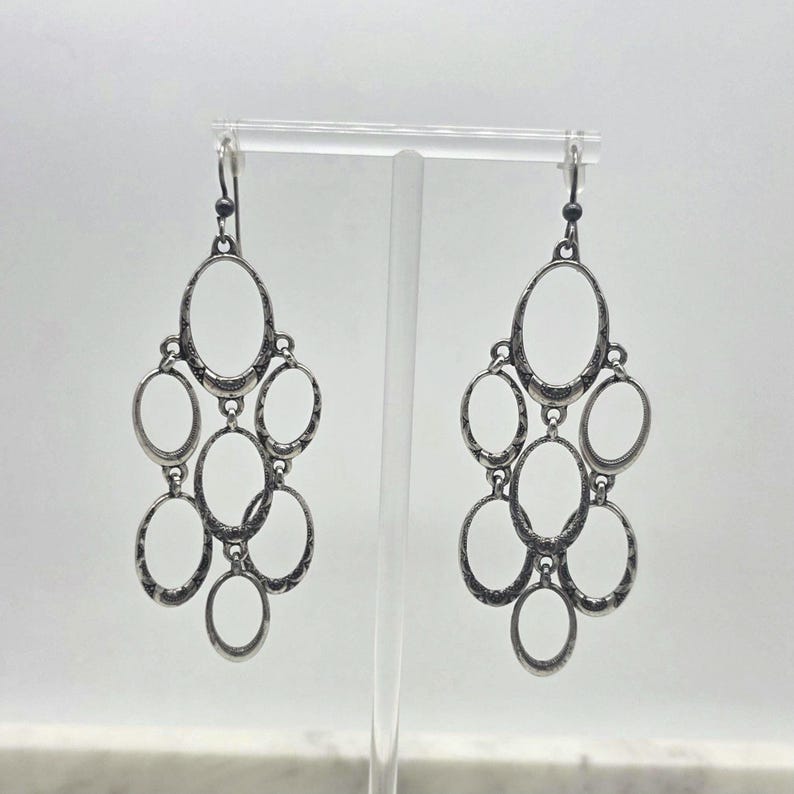 May include: A pair of silver-toned chandelier earrings. Each earring features multiple oval-shaped frames connected in a cascading design. The earrings are displayed against a white background, showcasing their intricate details and vintage style.