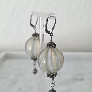 May include: A pair of dangle earrings with a vintage aesthetic. Each earring features a round, ribbed, clear glass orb with vertical white stripes. The orbs are suspended from black metal hooks and adorned with a small, dark gray pearl at the bottom.