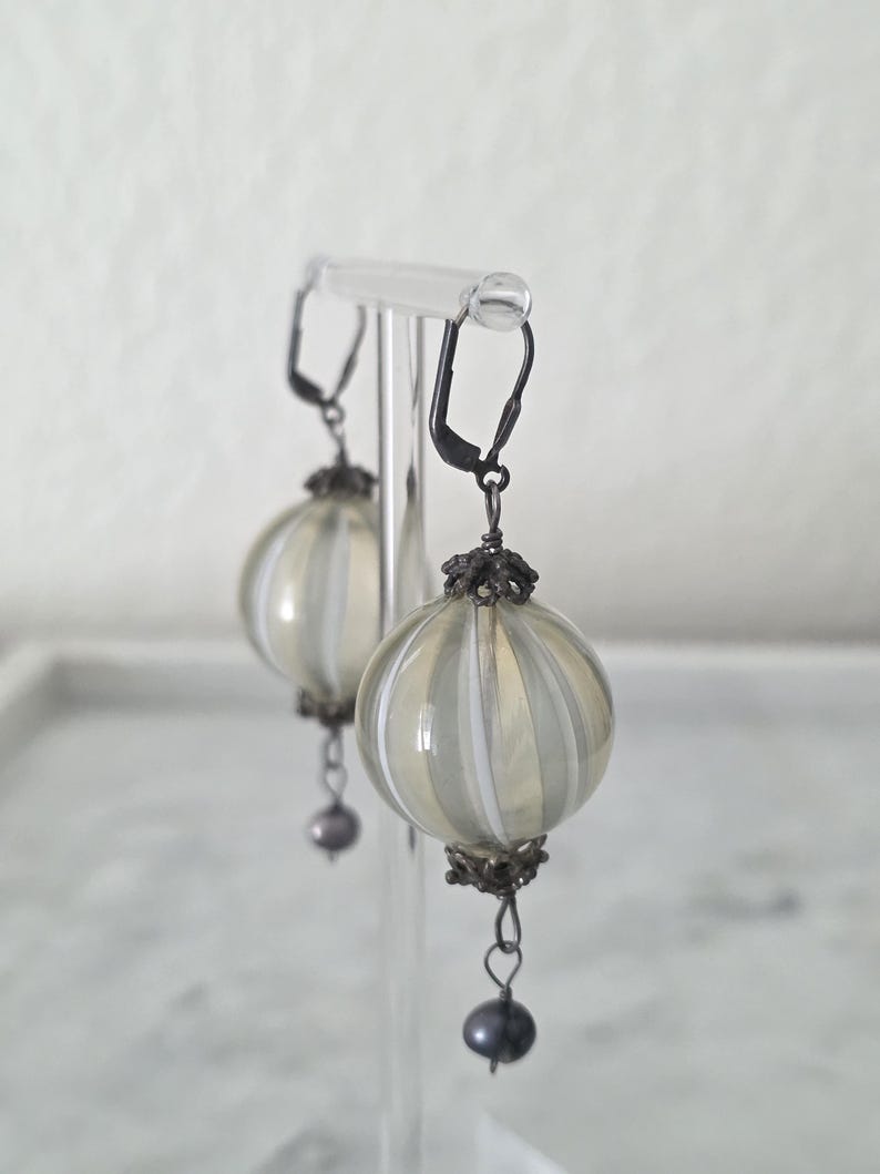 May include: A pair of dangle earrings with a vintage aesthetic. Each earring features a round, ribbed, clear glass orb with vertical white stripes. The orbs are suspended from black metal hooks and adorned with a small, dark gray pearl at the bottom.