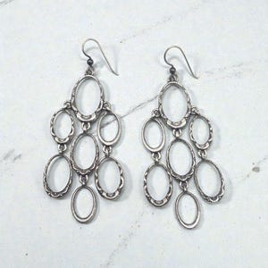 May include: A pair of silver chandelier earrings. Each earring features multiple oval-shaped frames connected in a cascading design. The frames have a decorative, textured edge and are suspended from a simple hook.
