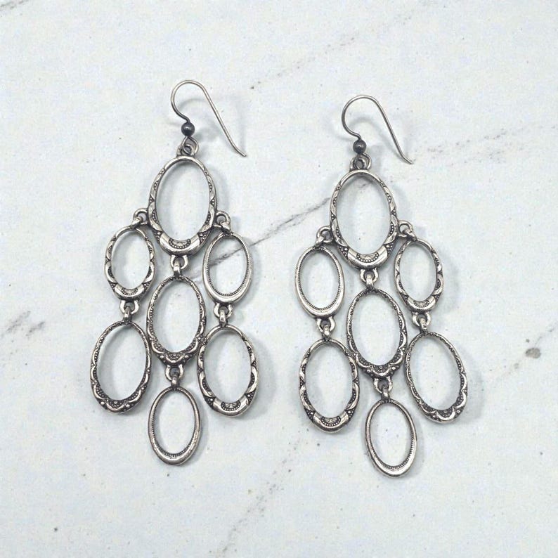 May include: A pair of silver chandelier earrings. Each earring features multiple oval-shaped frames connected in a cascading design. The frames have a decorative, textured edge and are suspended from a simple hook.