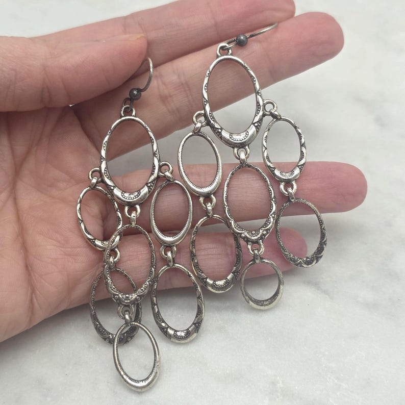 May include: Silver chandelier earrings with multiple oval-shaped links. The earrings have a vintage, ornate design with decorative detailing on the links. The earrings are suspended from a small hook.