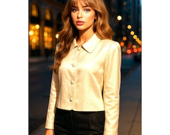 May include: A cream-colored, button-down jacket with a rounded collar. The jacket has long sleeves and is paired with black pants. The background features a blurred city street scene with lights.