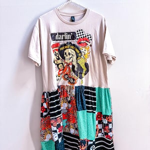 May include: A beige t-shirt dress featuring a graphic print of a skull wearing a hat with the text 'FUCK WHAT THEY THINK BE YOU' and 'darlin''. The skirt is a patchwork design with various patterns including leopard print, black and white stripes, flames, flowers, and green textures.