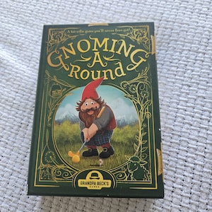 May include: A rectangular board game box titled Gnoming A Round by Grandpa Beck's Games. The dark green cover features a gold-outlined illustration of a gnome playing golf in a grassy field with mountains in the background and decorative floral accents.