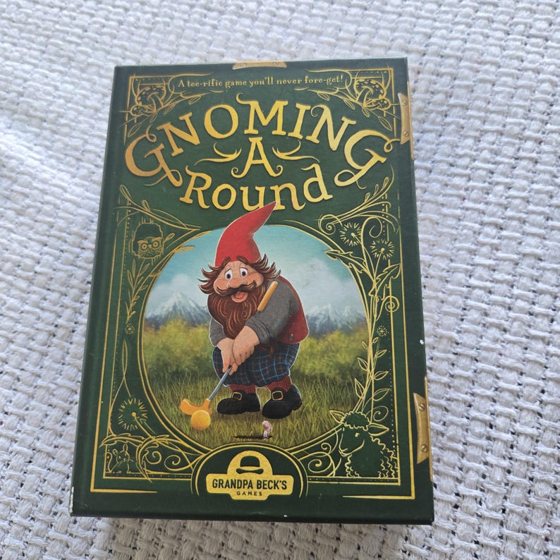 May include: A rectangular board game box titled Gnoming A Round by Grandpa Beck's Games. The dark green cover features a gold-outlined illustration of a gnome playing golf in a grassy field with mountains in the background and decorative floral accents.