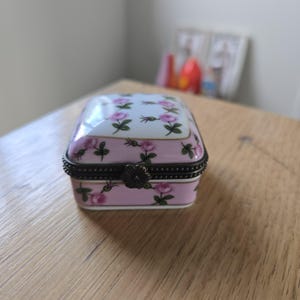 Może przedstawiać: Kwadratowe pudełko na drobiazgi z porcelany z białym wieczkiem i różową podstawą, ozdobione wzorem małych różowych róż i zielonych liści. Pudełko posiada ciemne metalowe zapięcie na zawiasach z klamrą w kształcie kwiatka z przodu.
