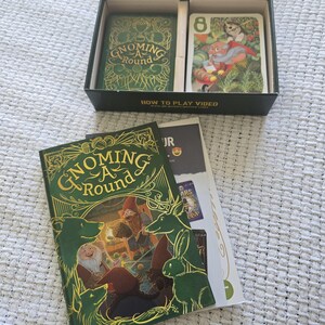 May include: A Gnoming A Round card game set featuring a black box with two card compartments, a rulebook, and a game manual. The box lid and manual cover display a green and gold illustration of gnomes, a bear, a deer, and a rabbit.
