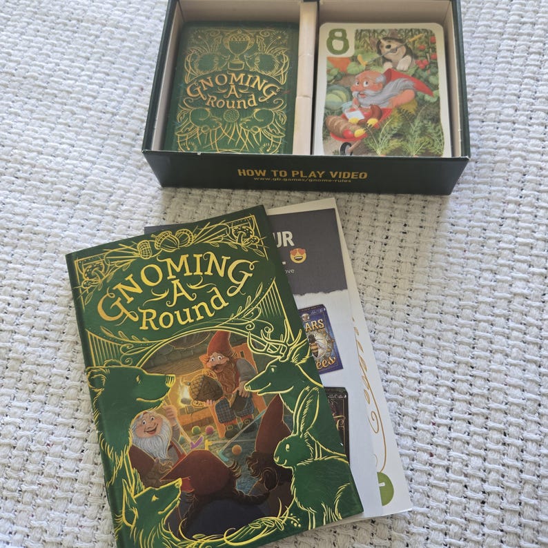 May include: A Gnoming A Round card game set featuring a black box with two card compartments, a rulebook, and a game manual. The box lid and manual cover display a green and gold illustration of gnomes, a bear, a deer, and a rabbit.