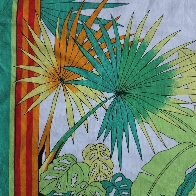 May include: A white textile with a tropical leaf pattern. The design features large green, yellow, and orange palm leaves and other tropical foliage. A vertical stripe with red, orange, and green stripes borders the left side.
