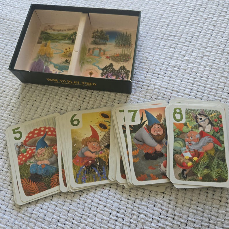 May include: A board game box with a divided interior featuring landscape illustrations and a set of playing cards numbered five through eight. Each card displays colorful artwork of gnomes in garden settings, with the text HOW TO PLAY VIDEO.