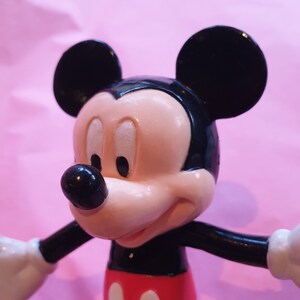 Peut inclure: Gros plan sur une figurine de Mickey Mouse. La figurine a une t&ecirc;te noire avec de grandes oreilles rondes, un visage rose et un nez noir. Il porte un short rouge avec deux cercles blancs et des bras noirs. Le fond est rose uni.