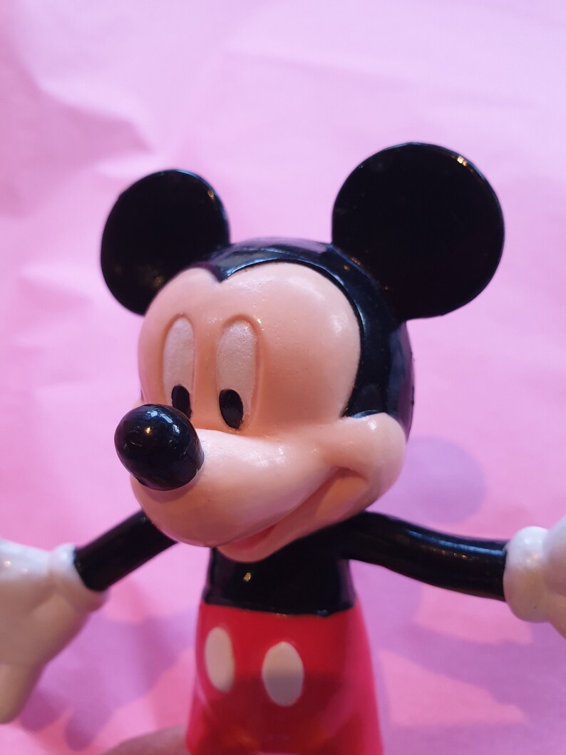 Peut inclure: Gros plan sur une figurine de Mickey Mouse. La figurine a une t&ecirc;te noire avec de grandes oreilles rondes, un visage rose et un nez noir. Il porte un short rouge avec deux cercles blancs et des bras noirs. Le fond est rose uni.