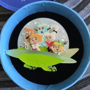 May include: A colourful brooch featuring the Flintstones family and their robot friend in a green spaceship against a white moon backdrop. The brooch is set in a blue circular container with a black interior.