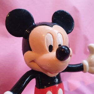 Peut inclure: Gros plan d'une figurine de Mickey Mouse. La figurine a des oreilles noires, un nez noir et un visage couleur p&ecirc;che. Mickey porte un haut noir, un short rouge avec des boutons blancs et des gants blancs. Le fond est rose uni.