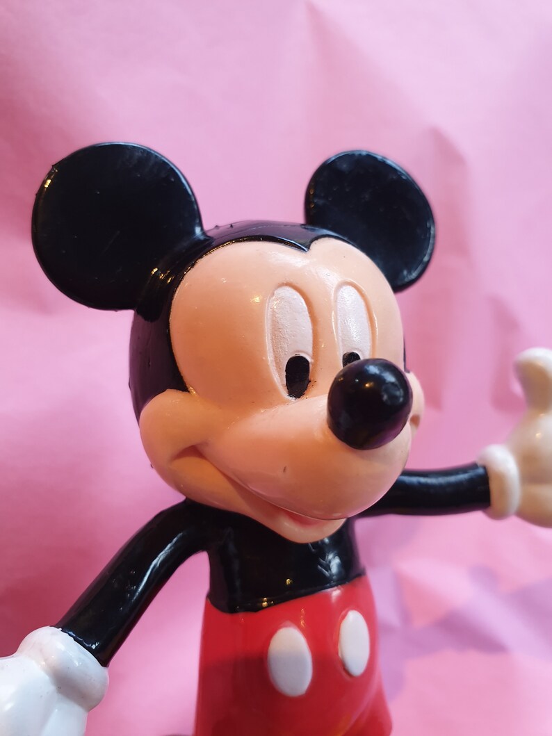 Peut inclure: Gros plan d'une figurine de Mickey Mouse. La figurine a des oreilles noires, un nez noir et un visage couleur p&ecirc;che. Mickey porte un haut noir, un short rouge avec des boutons blancs et des gants blancs. Le fond est rose uni.