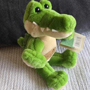 May include: A plush crocodile toy featuring bright green fur, a cream-coloured belly, and large black plastic eyes. The stuffed animal has a wide snout with visible white fabric teeth and a small tan patch on its chest, accompanied by a paper tag.
