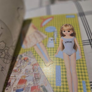 May include: A paper doll with blonde hair and a light blue swimsuit with the word "LICCA" printed on it. The doll is on a yellow and green patterned background with other cut-out doll parts. The background includes a drawing of a city.