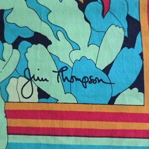 May include: A colorful textile featuring a vibrant floral pattern in shades of teal, blue, and green. The design includes the signature "Jim Thompson" in black script. The border has horizontal stripes of orange, red, and blue.