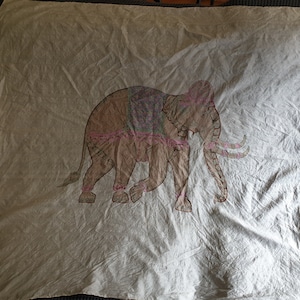 May include: A beige textile with a detailed illustration of an elephant. The elephant is depicted in shades of brown and pink, with decorative accents on its back and legs. The textile has a slightly wrinkled appearance.