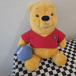 Puede incluir: Un peluche de Winnie the Pooh, con pelaje amarillo, una camisa roja y una olla azul. El juguete está sentado sobre una superficie a cuadros blancos y negros. La camisa tiene un pequeño parche de "Pooh".