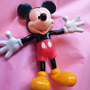 Peut inclure: Figurine de Mickey Mouse avec une t&ecirc;te et des oreilles noires, un visage beige et un nez noir. Il porte un short rouge avec des boutons blancs, des chaussures jaunes et des gants blancs. La figurine est pos&eacute;e sur un fond rose, les bras tendus.