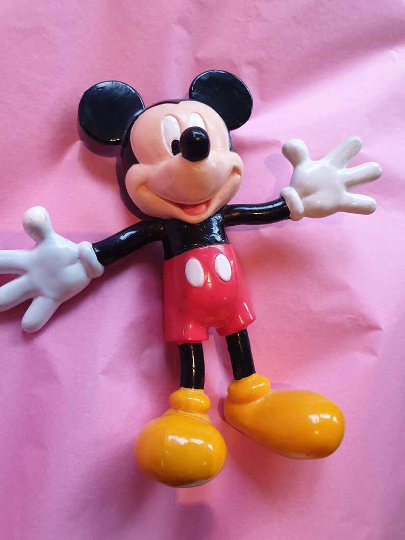 Peut inclure: Figurine de Mickey Mouse avec une t&ecirc;te et des oreilles noires, un visage beige et un nez noir. Il porte un short rouge avec des boutons blancs, des chaussures jaunes et des gants blancs. La figurine est pos&eacute;e sur un fond rose, les bras tendus.