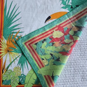 May include: White square scarf with a tropical design. The scarf features a toucan, palm leaves, and floral patterns in green, orange, and red. The border has red and orange stripes. The corner has a signature in cursive.