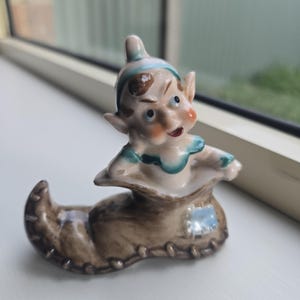 Peut inclure: Figurine vintage en céramique d'un elfe fantaisiste sortant d'une botte marron. L'elfe a la peau claire, les joues roses et une expression espiègle. La botte a un patch bleu et est détaillée avec des coutures.