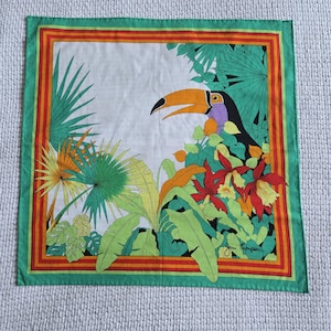 May include: Square silk scarf with a toucan and tropical foliage design. The scarf features a toucan with a large orange beak, surrounded by green, yellow, and orange leaves and flowers. The border is green with orange and red stripes.