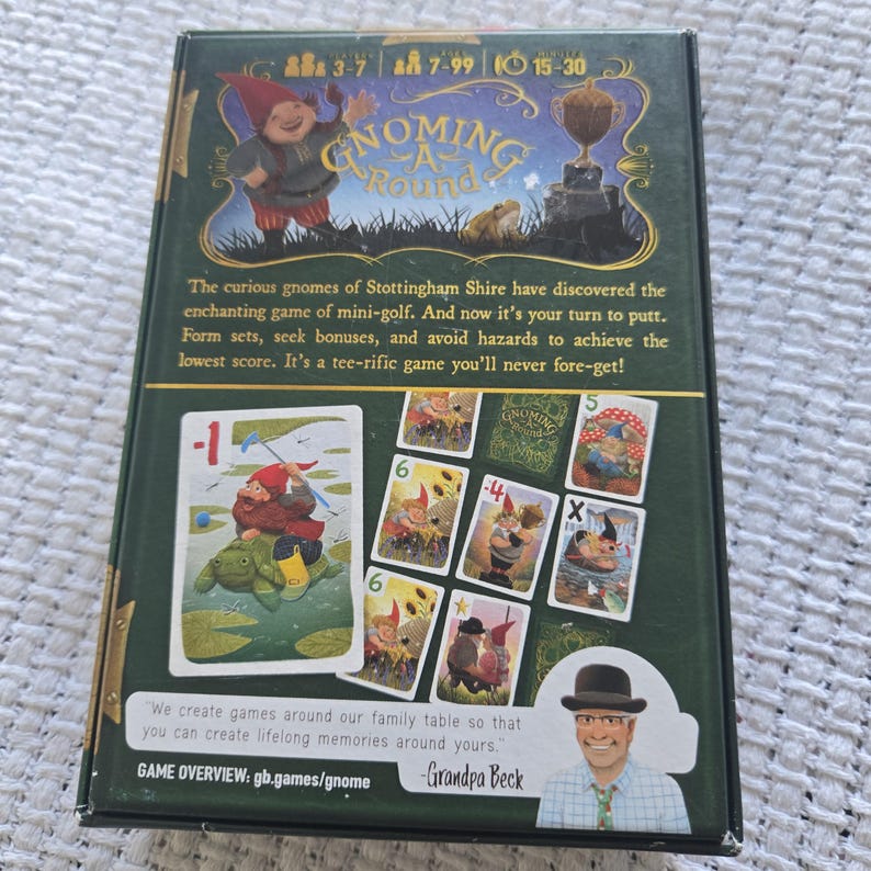 May include: A rectangular board game box titled Gnoming A Round with a dark green background. The box features illustrations of gnomes, a trophy, and playing cards with numerical values. Text includes game details, rules, and a quote from Grandpa Beck.