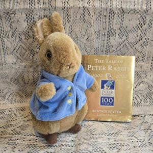 Puede incluir: Un peluche de Peter Rabbit con una chaqueta azul con botones amarillos, junto a un libro dorado titulado "The Tale of Peter Rabbit" de Beatrix Potter. El libro conmemora los 100 años de Peter Rabbit.