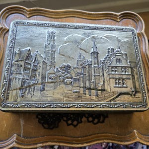 Puede incluir: Caja metálica rectangular con un diseño de paisaje urbano en relieve. La caja presenta una combinación de colores oro y plata, con un borde decorativo. El diseño en relieve representa edificios, una torre alta y nubes.