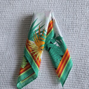May include: A folded white silk scarf with a tropical leaf design. The scarf features green, orange, and yellow palm fronds and a green border with orange and red stripes. The scarf is folded in a triangle shape.