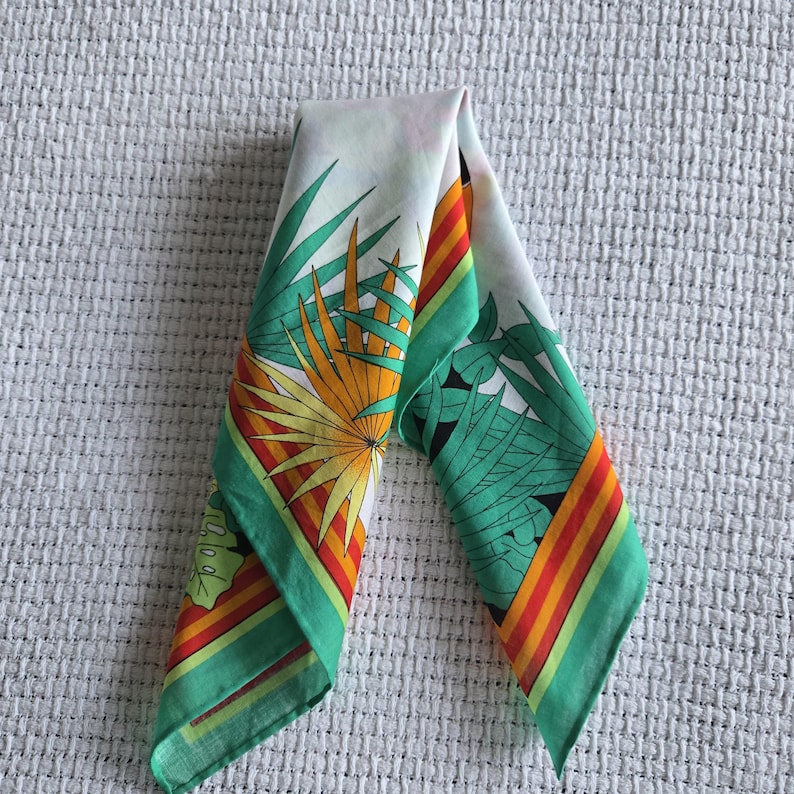 May include: A folded white silk scarf with a tropical leaf design. The scarf features green, orange, and yellow palm fronds and a green border with orange and red stripes. The scarf is folded in a triangle shape.