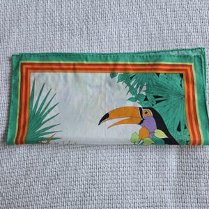 May include: A folded square scarf with a toucan design. The scarf features a white background with a toucan, green leaves, and a green, orange, and red border. The toucan has a large orange beak and purple accents.