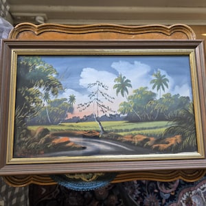 May include: A framed painting depicting a tropical landscape with palm trees, lush greenery, and a winding path. The sky is a vibrant blue with fluffy white clouds. The painting is done in a realistic style with a focus on detail.