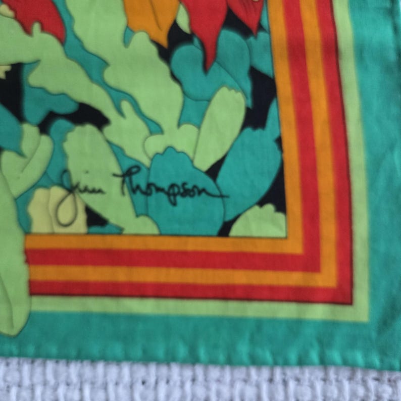 May include: A square silk scarf with a colorful floral design. The scarf features a teal border with orange, red, and yellow stripes. The center has green, teal, and red floral shapes with the signature "Jim Thompson" in black.