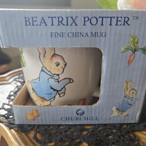 以下が含まれることがあります： オリジナルボックスに入った、ビアトリクス・ポターのデザインが施された上質な磁器マグカップ。マグカップには、青いジャケットを着たウサギの魅力的なイラストが描かれています。箱は水色で、「BEATRIX POTTER」と「FINE CHINA MUG」の文字が書かれています。