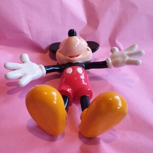 Peut inclure: Figurine de Mickey Mouse dans une pose d&eacute;tendue, les bras tendus et les jambes &eacute;cart&eacute;es. La figurine pr&eacute;sente un corps noir, un short rouge, des chaussures jaunes et des gants blancs. Le fond est une surface rose textur&eacute;e.
