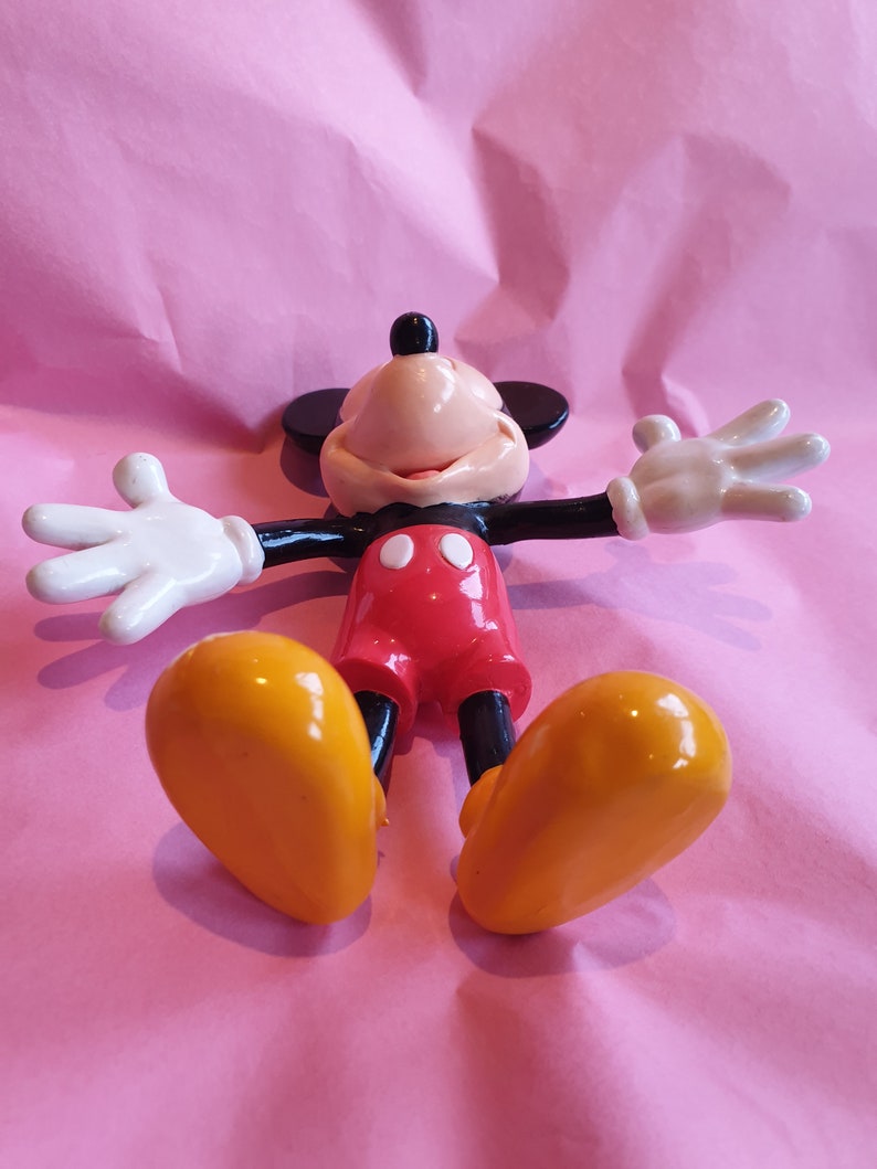 Peut inclure: Figurine de Mickey Mouse dans une pose d&eacute;tendue, les bras tendus et les jambes &eacute;cart&eacute;es. La figurine pr&eacute;sente un corps noir, un short rouge, des chaussures jaunes et des gants blancs. Le fond est une surface rose textur&eacute;e.