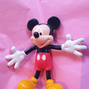 Peut inclure: Figurine de Mickey Mouse avec des oreilles noires, une t&ecirc;te noire et un visage beige. Il porte un short rouge, des chaussures jaunes et des gants blancs. La figurine est pos&eacute;e avec les bras tendus sur un fond rose.