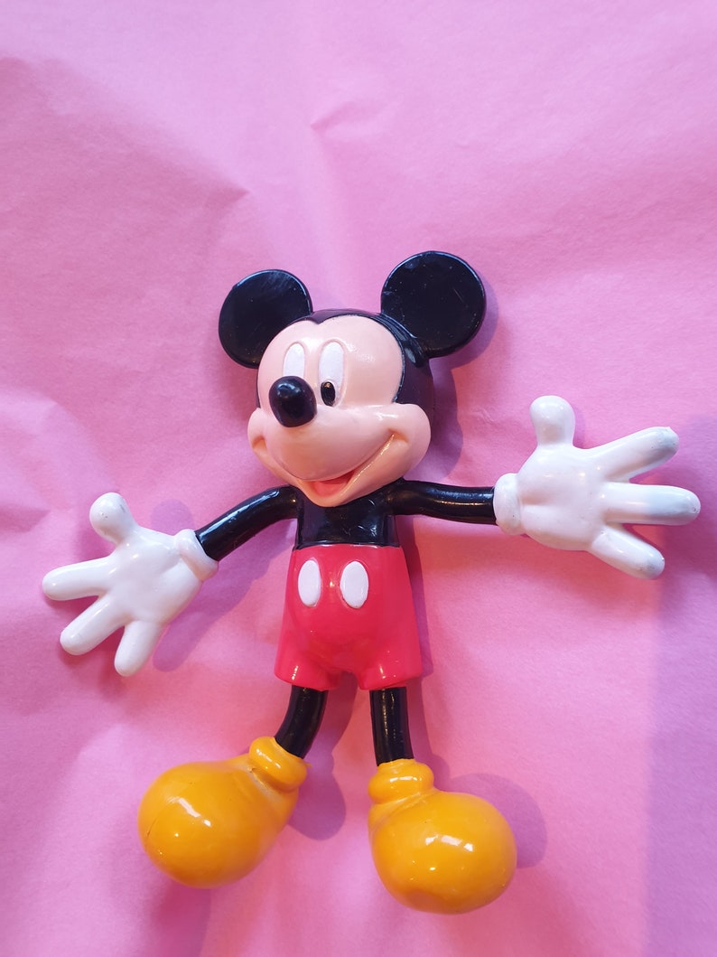Peut inclure: Figurine de Mickey Mouse avec des oreilles noires, une t&ecirc;te noire et un visage beige. Il porte un short rouge, des chaussures jaunes et des gants blancs. La figurine est pos&eacute;e avec les bras tendus sur un fond rose.