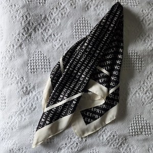 May include: Black and white square scarf with the word "PRINGLE" repeated diagonally across the fabric. The scarf has a white border and is folded, displaying the brand name prominently. The scarf is resting on a white, textured surface.