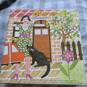 Puede incluir: Una caja vintage de Stencil Art 2 de Tuppertoy. La caja presenta una colorida ilustración de una mujer con un vestido floral, un gato negro y un coche. El texto de la caja dice: "Una herramienta de aprendizaje creativa y educativa para crecer con tu hijo".