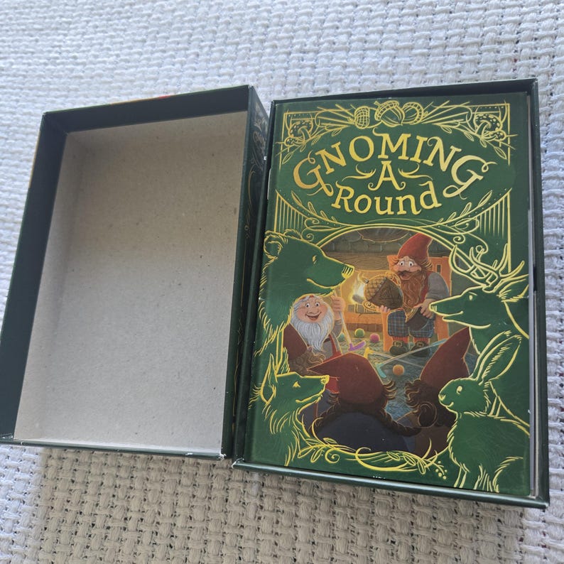 May include: A rectangular game box with an open lid. The cover features the text Gnoming A Round in gold lettering, surrounded by green illustrations of a gnome, a bear, a deer, and a rabbit in a forest setting with a fireplace background.