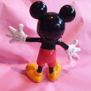 Peut inclure: Figurine de Mickey Mouse, vue de dos, sur une surface rose. La figurine a une t&ecirc;te noire avec de grandes oreilles rondes, des gants blancs, un short rouge et des chaussures jaunes. Les bras de la figurine sont tendus.