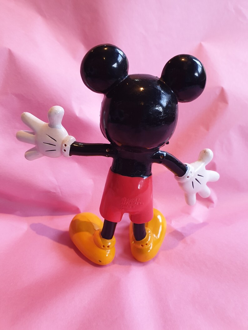 Peut inclure: Figurine de Mickey Mouse, vue de dos, sur une surface rose. La figurine a une t&ecirc;te noire avec de grandes oreilles rondes, des gants blancs, un short rouge et des chaussures jaunes. Les bras de la figurine sont tendus.