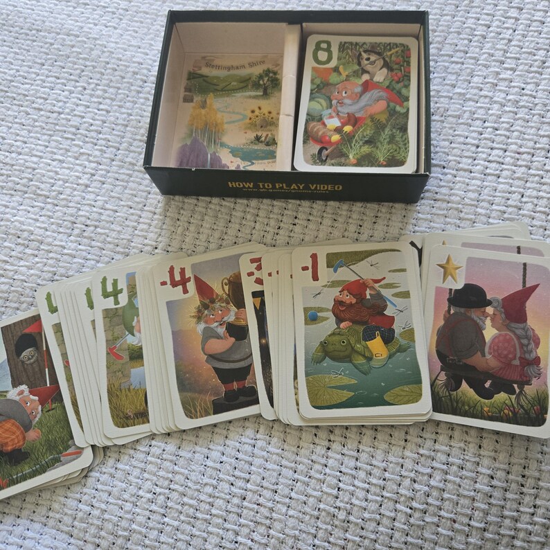 May include: A card game set featuring a black box with the text HOW TO PLAY VIDEO and a deck of cards. The cards display colorful illustrations of gnomes in various outdoor settings, with numbers and symbols like 8, 4, -4, -3, -1, and a star.
